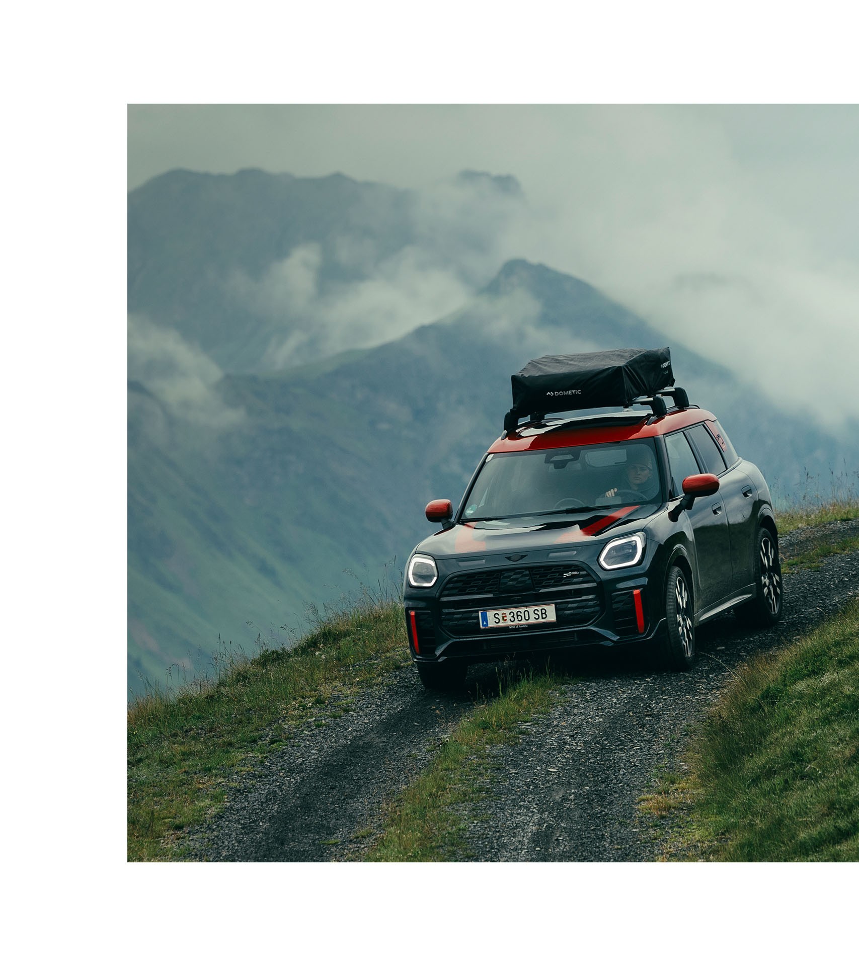 The new MINI John Cooper Works Countryman ALL4 with roof rack pictured driving around a corner on an unpaved mountain road, with a steep drop-off and misty mountain views in the background.